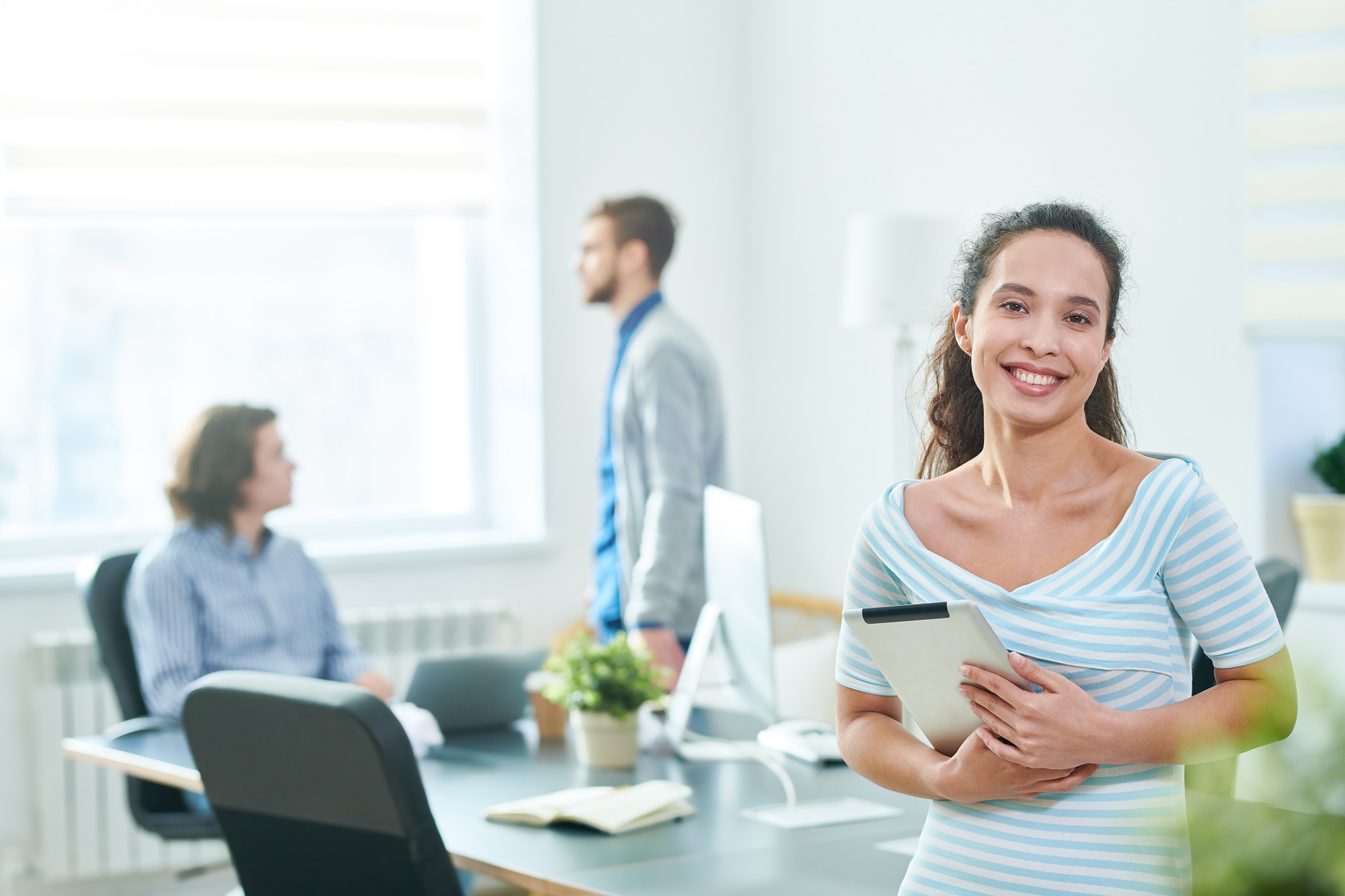 Happy successful stylish young Hispanic female manager embracing tablet and looking at camera cheerfully while her colleagues discussing project in background